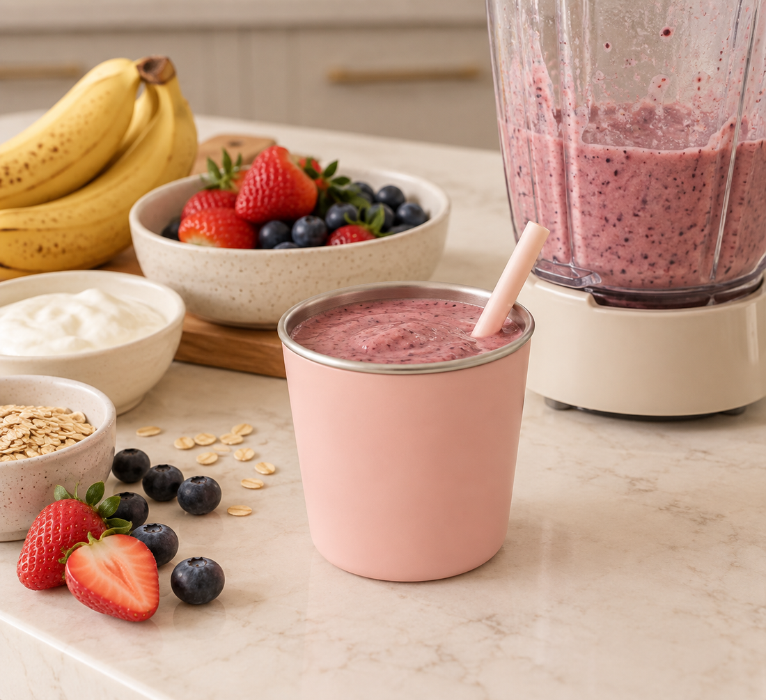 BRAI + CO's Pink stainless steel toddler cup filled with a berry smoothie beside a blender, fresh strawberries, blueberries, banana, Greek yogurt and oats.
